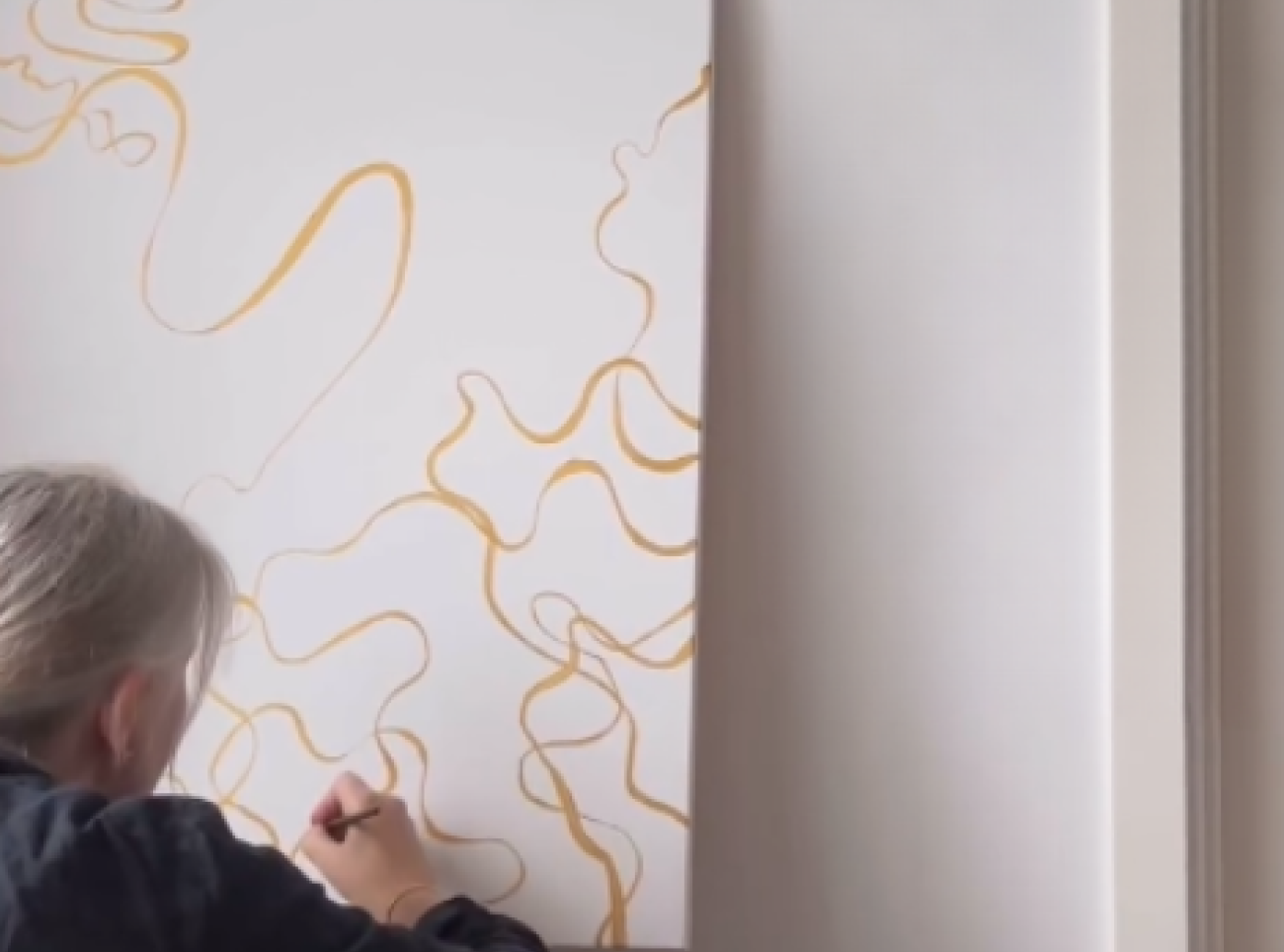 Karin Margarita Frei, wearing dark clothing, is seated on the floor drawing abstract orange lines on a white canvas resting against a wall.